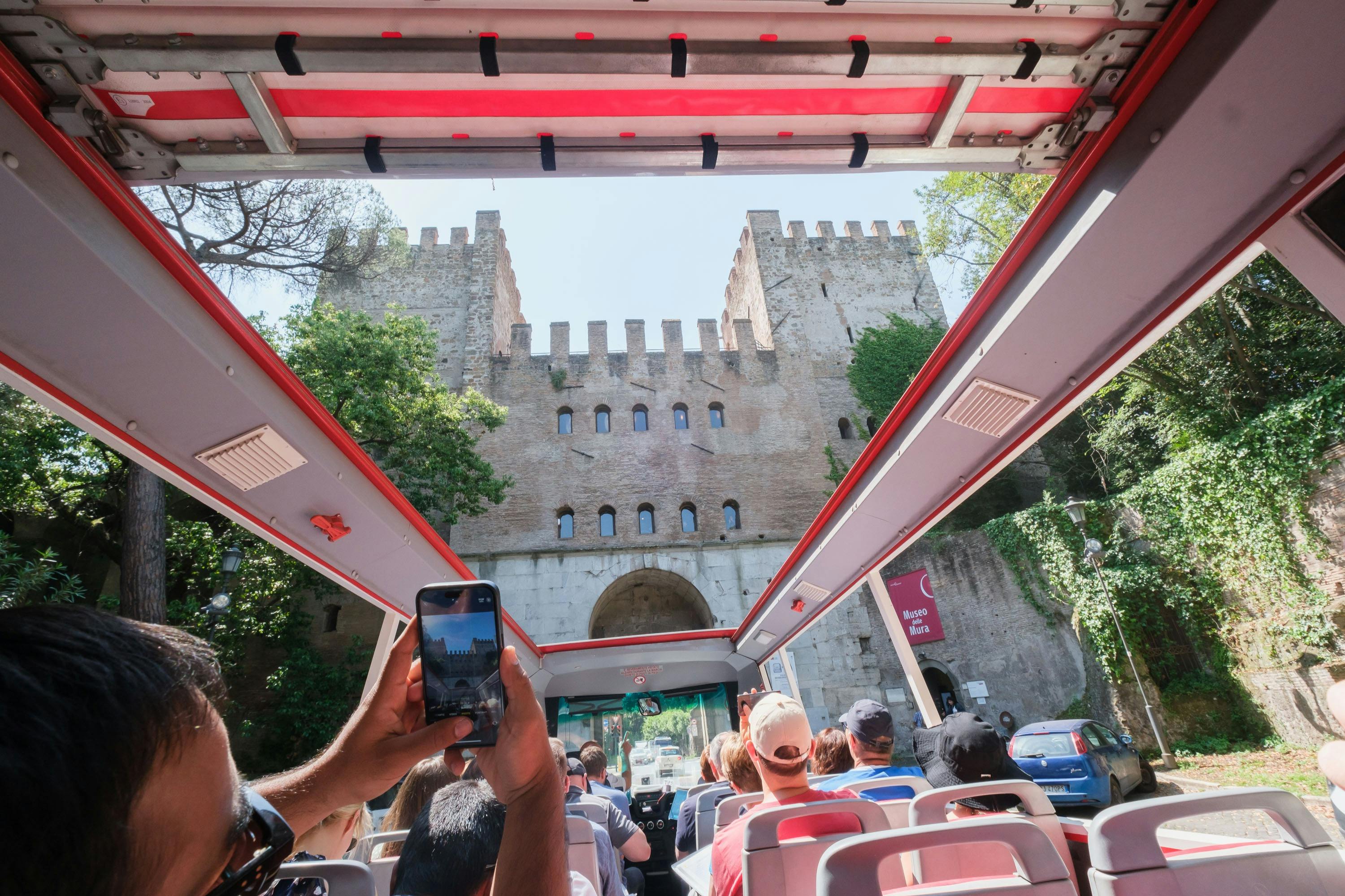 Vue depuis un bus touristique à toit ouvert s'approchant d'une porte en pierre aux allures de château, avec des passagers en train de prendre des photos.