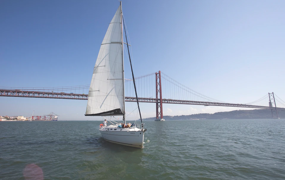 A sailboat with raised sails in the water near a large, red suspension bridge on a clear, sunny day.
