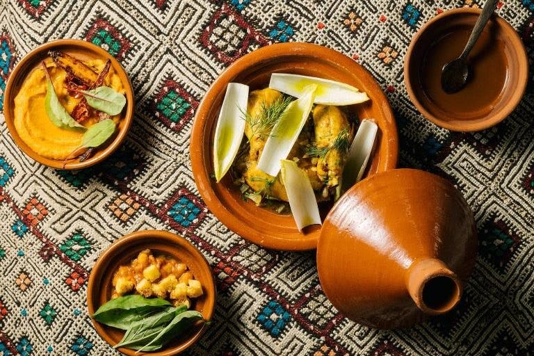 Earthy pottery dishes filled with various cooked foods, set on a patterned fabric. One dish is covered with a matching lid.