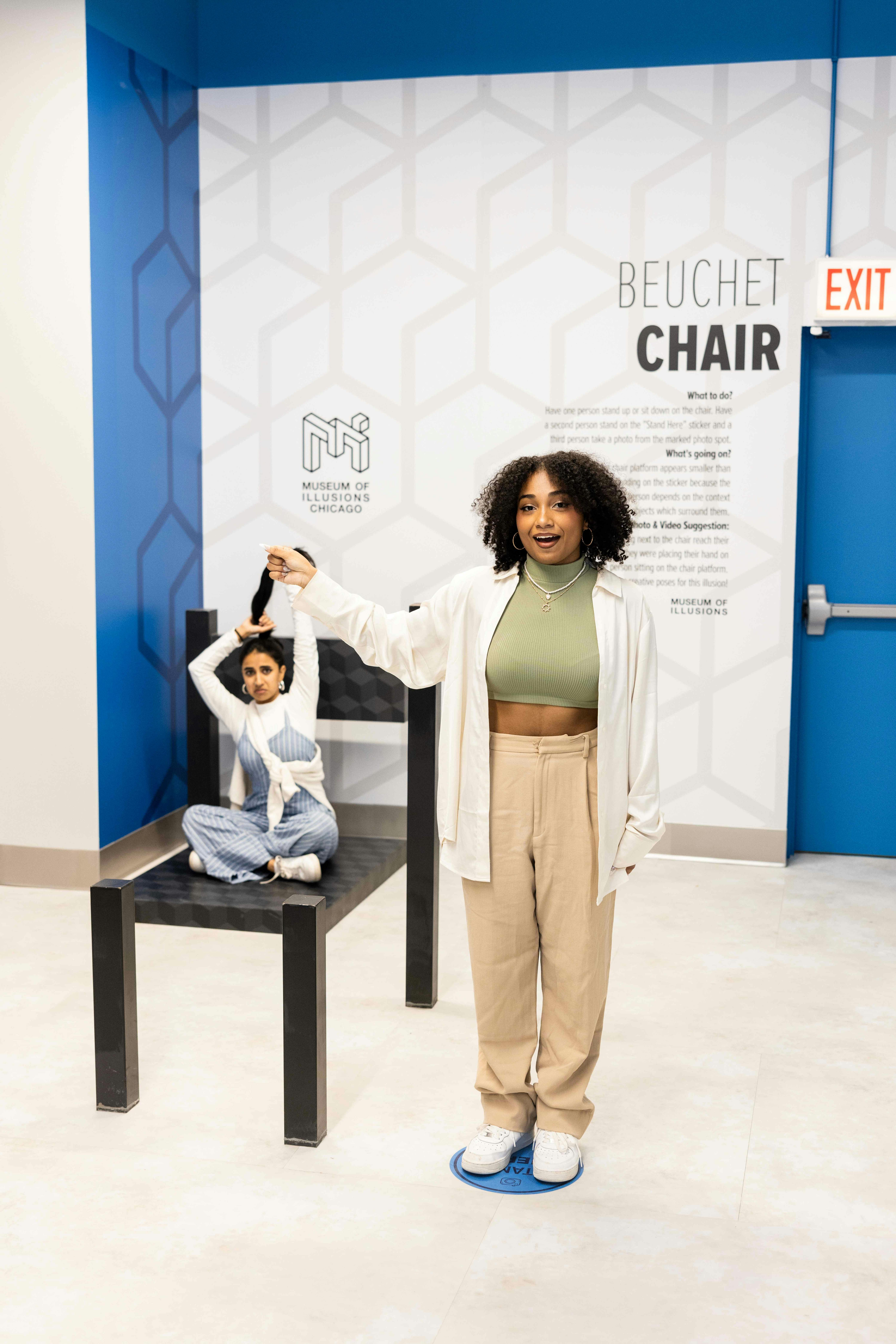 Two women in an illusion exhibit; one appears small on a chair prop, while the other stands in front, holding her hand above the chair.