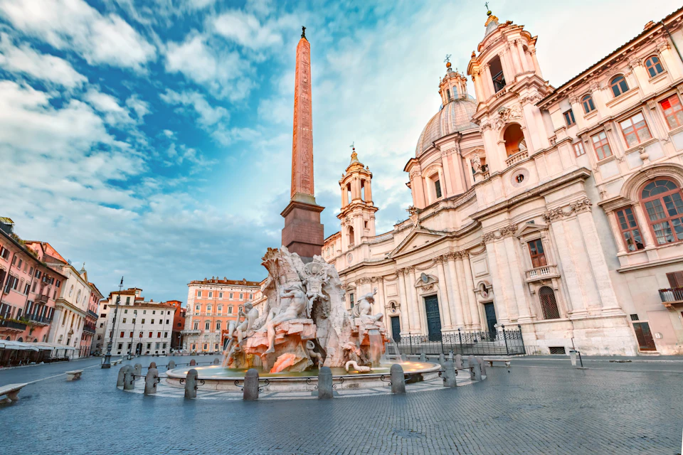 Historic square with a large, ornate fountain featuring statues and an obelisk, adjacent to a baroque-style building with domes.