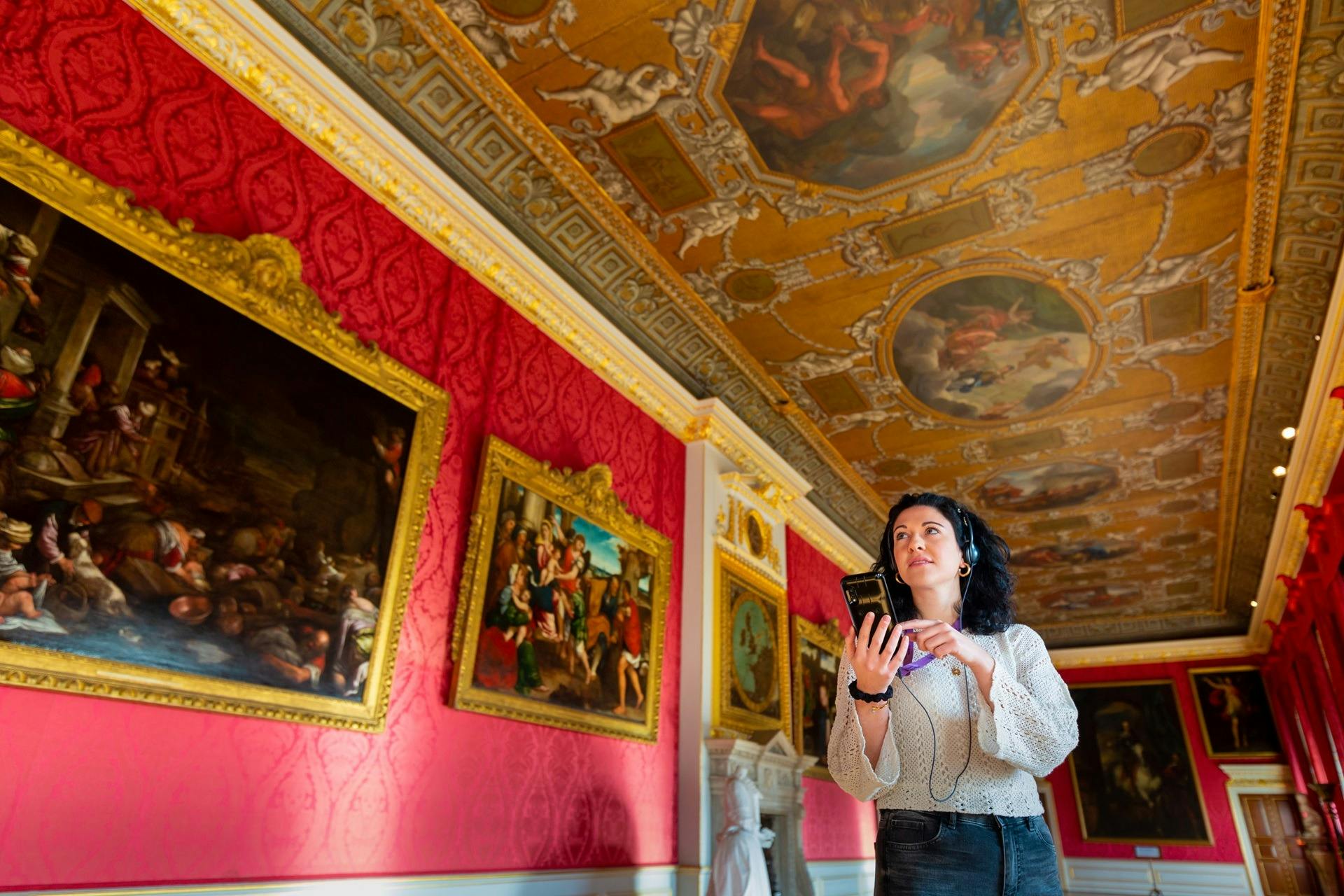 A woman using an audio guide device in an ornate room with red walls, large framed paintings, and a decorated ceiling.