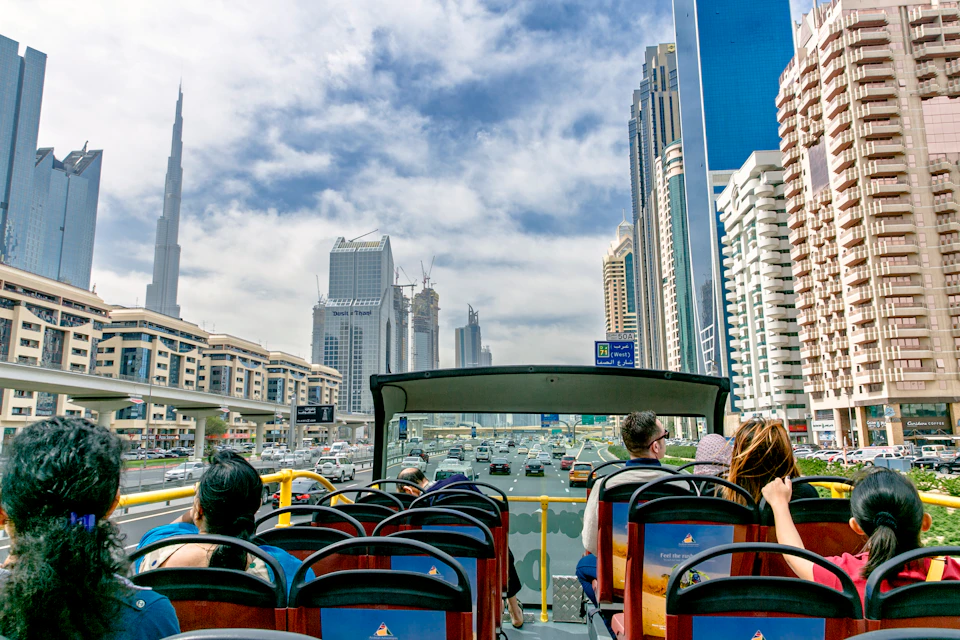 Passengers on an open-top tour bus view a cityscape with skyscrapers and highways under a partly cloudy sky.