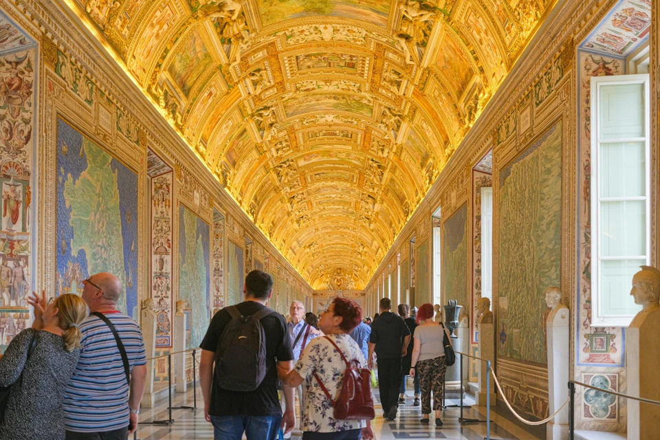 People walking through a long, ornate hallway with a gold-decorated ceiling and detailed wall artwork.