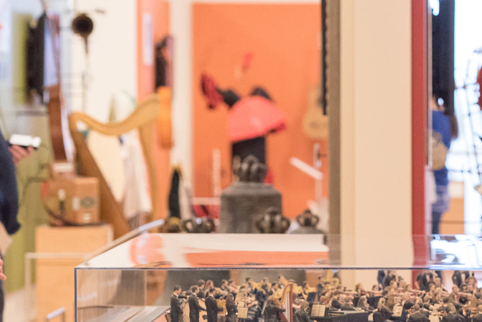 People observing a detailed miniature display in a museum exhibit, with various artifacts and musical instruments in the background.