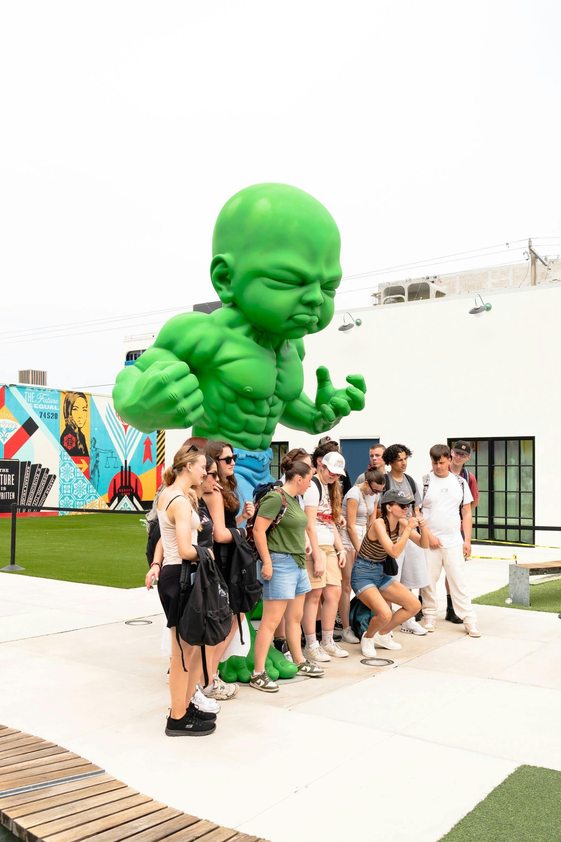 一群人站在户外市区的一个大型肌肉发达的绿色婴儿雕像前。