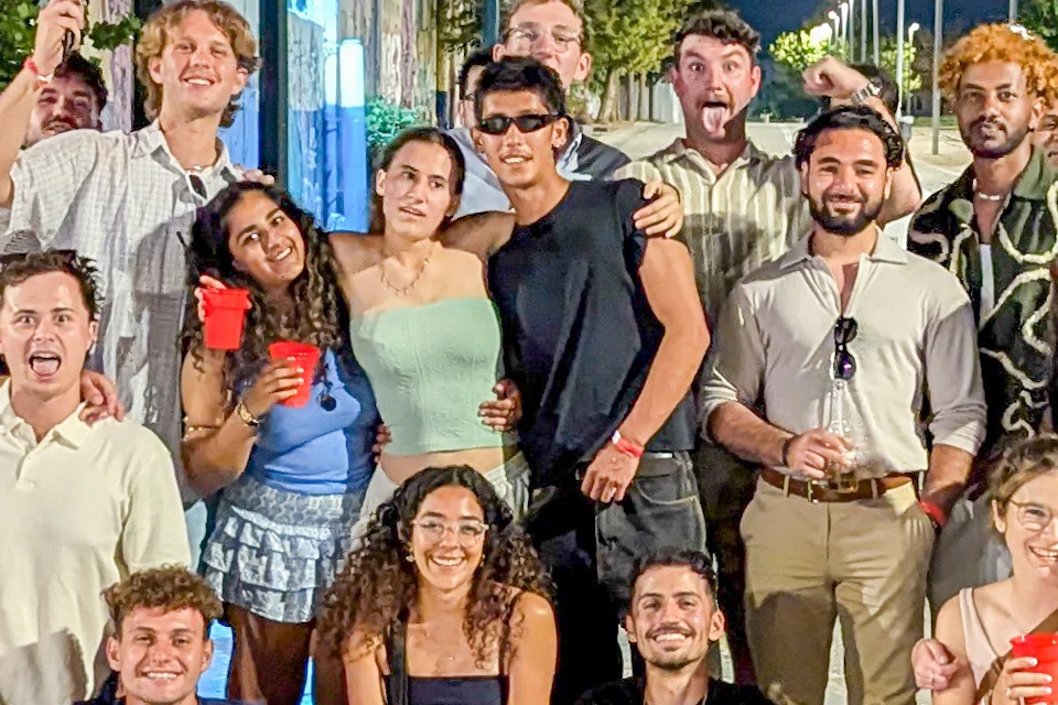 A group of people posing together outdoors at night in a well-lit area, some holding red cups, with trees and a street in the background.