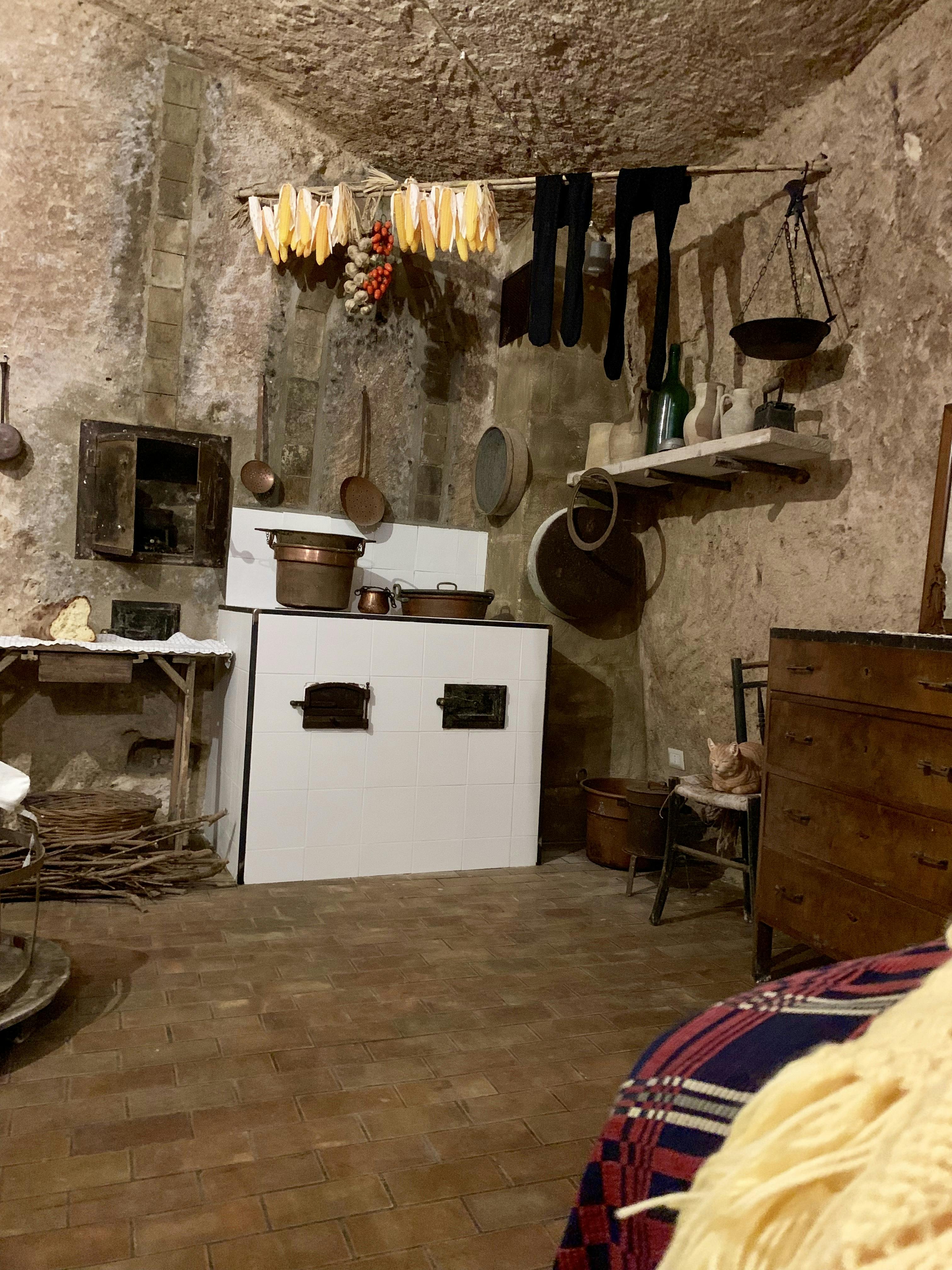 Una cucina rustica con pareti in pietra, una stufa a legna, erbe da essiccare, scaffali in legno e vari utensili da cucina.