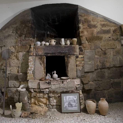 Ancient stone oven with various pottery items displayed on and near it, including jugs, bowls, and a framed photo.