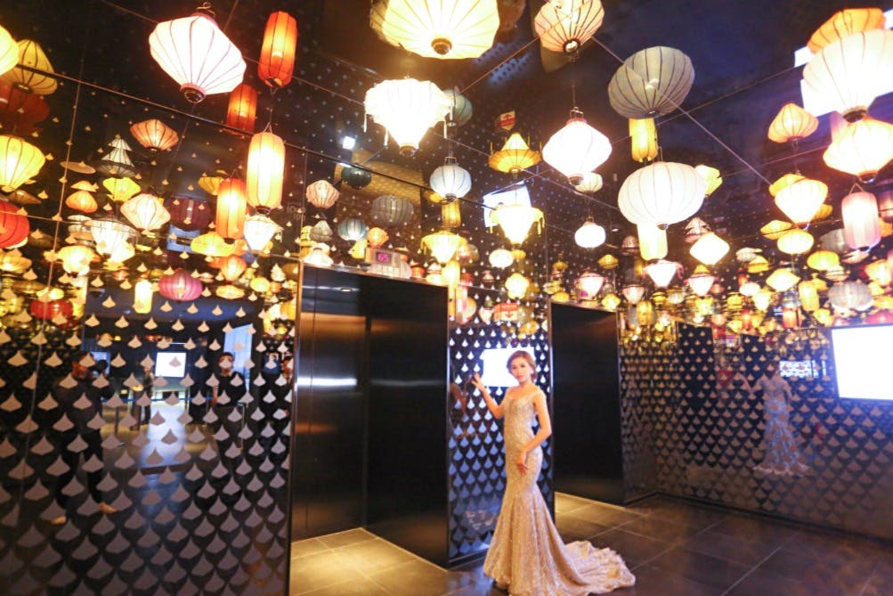 A woman in an evening gown stands in a mirrored room with numerous colorful hanging lanterns and two elevator doors.