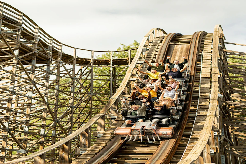 People riding a wooden rollercoaster, raising their hands as they descend a steep slope.