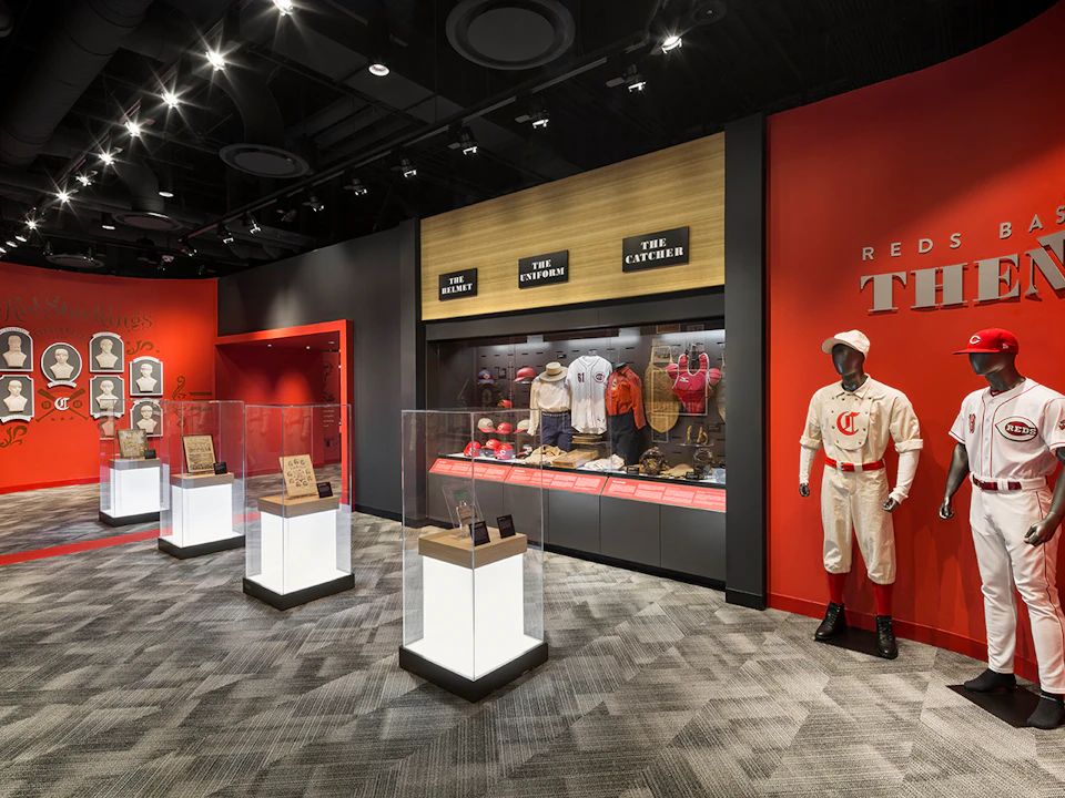 A museum exhibit displays vintage baseball uniforms, equipment, and memorabilia in glass cases, with a mannequin wearing an old-fashioned uniform.
