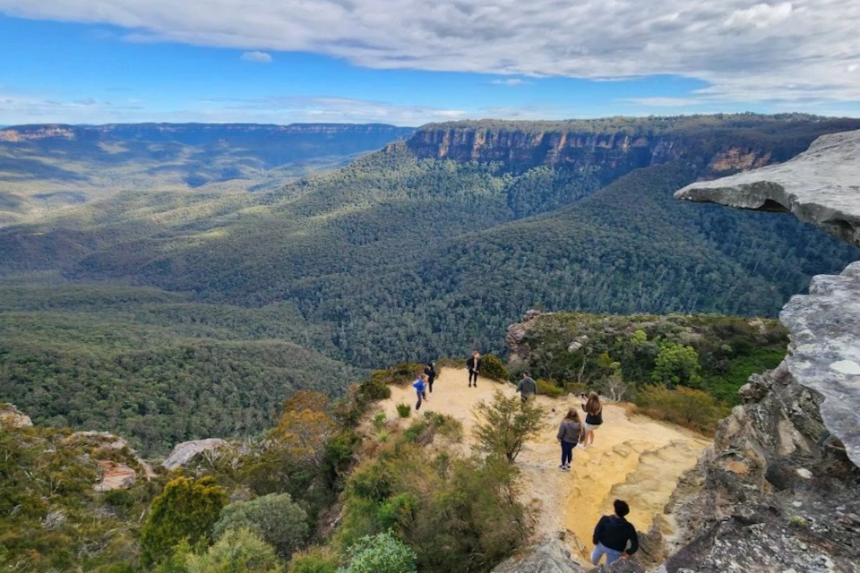 Several people stand on a cliff edge overlooking a vast, forested valley and distant mountains under a partly cloudy sky.