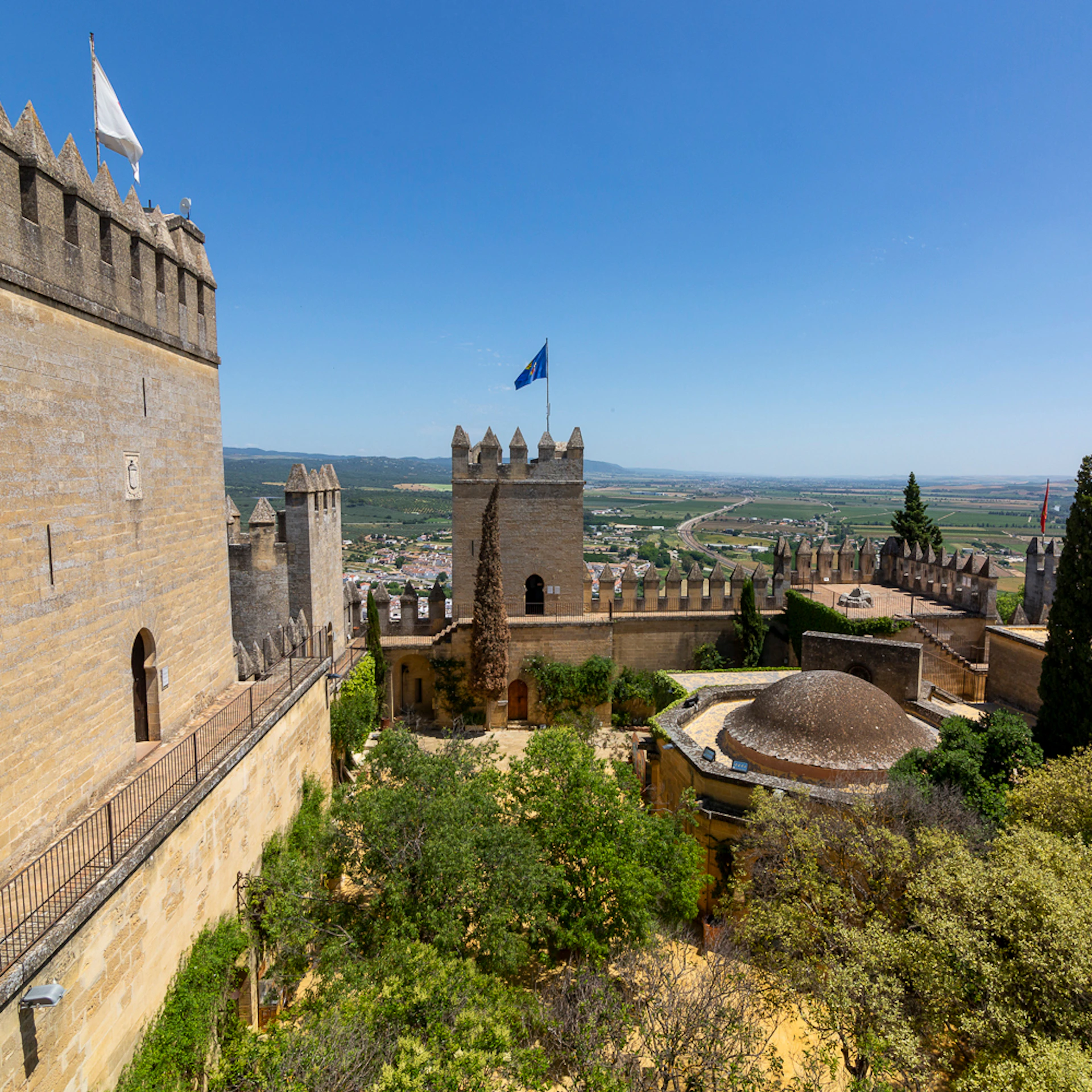 Stone fortress with crenellated walls and a flag on a central tower, surrounded by greenery and overlooking a vast rural landscape.