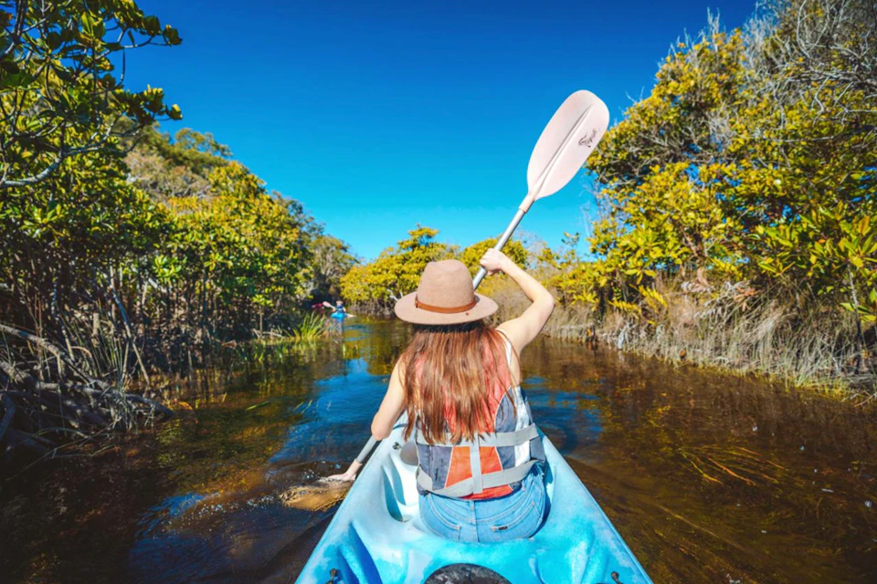 A person with a hat kayaking on a narrow waterway surrounded by dense vegetation under a clear blue sky.