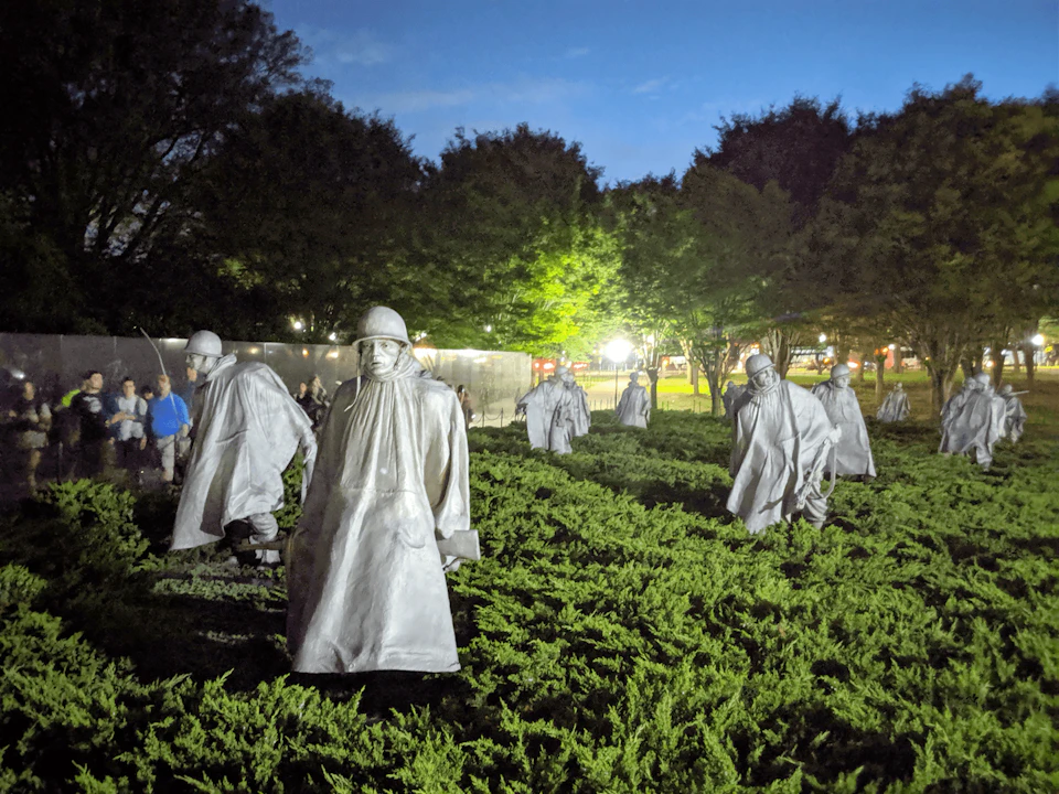 Statues of soldiers in raincoats stand among greenery, lit up at twilight with trees and blurred lights in the background.