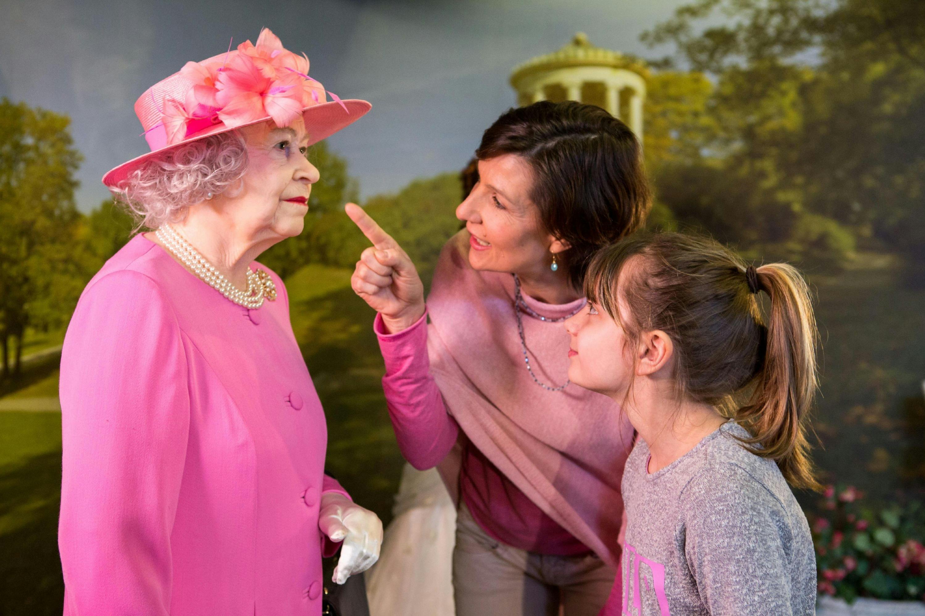A woman and a child interacting with a wax figure of a person dressed in a pink outfit and hat, with a scenic background.