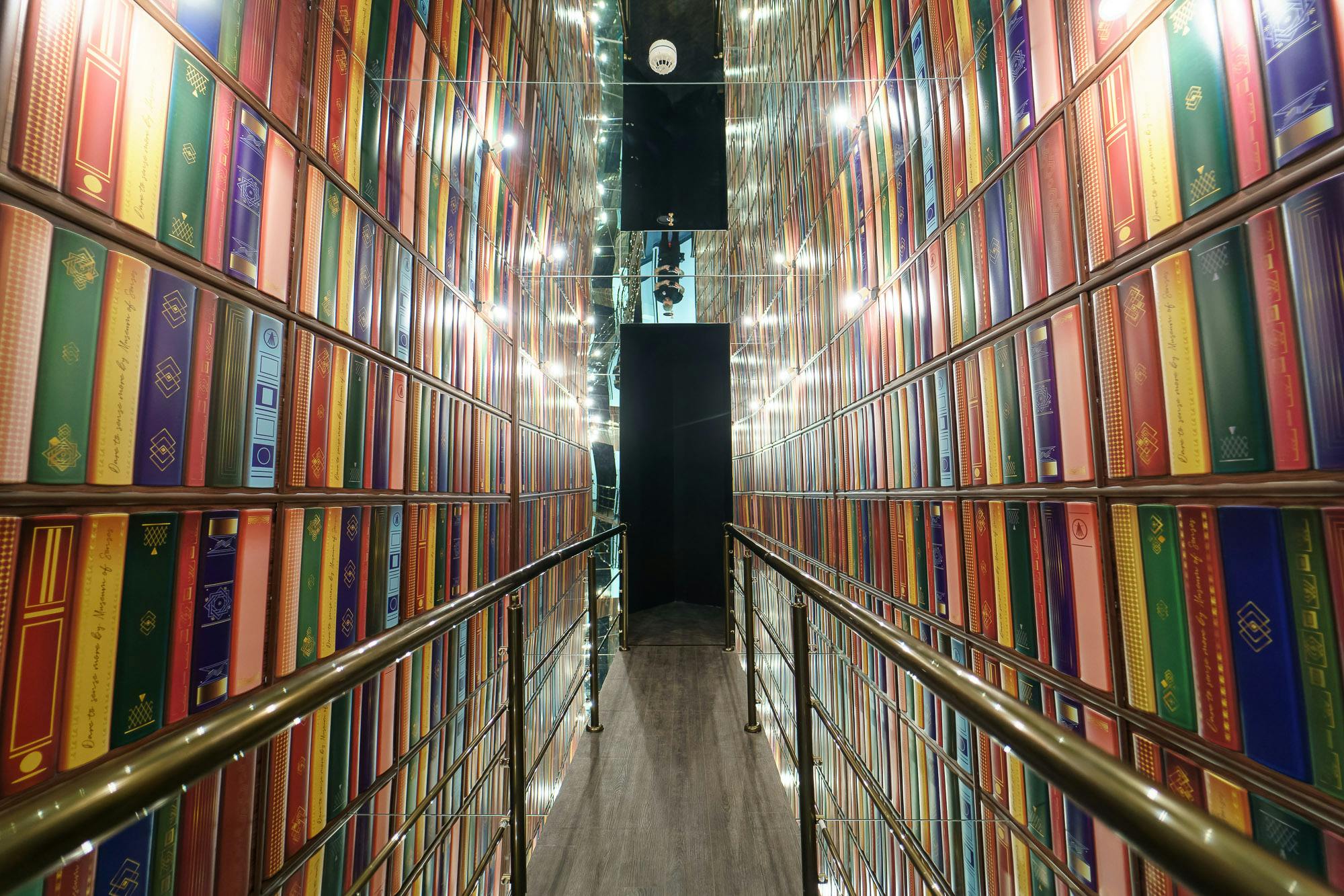 Narrow hallway with walls lined with colorful books, reflective ceiling, and metal railings on both sides.