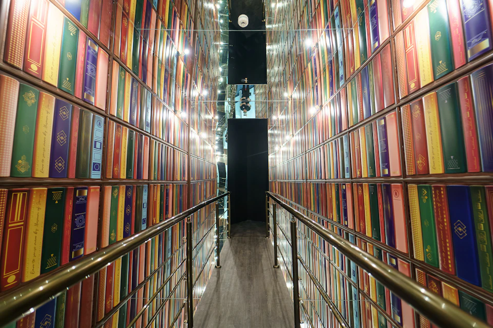 Narrow hallway with walls lined with colorful books, reflective ceiling, and metal railings on both sides.