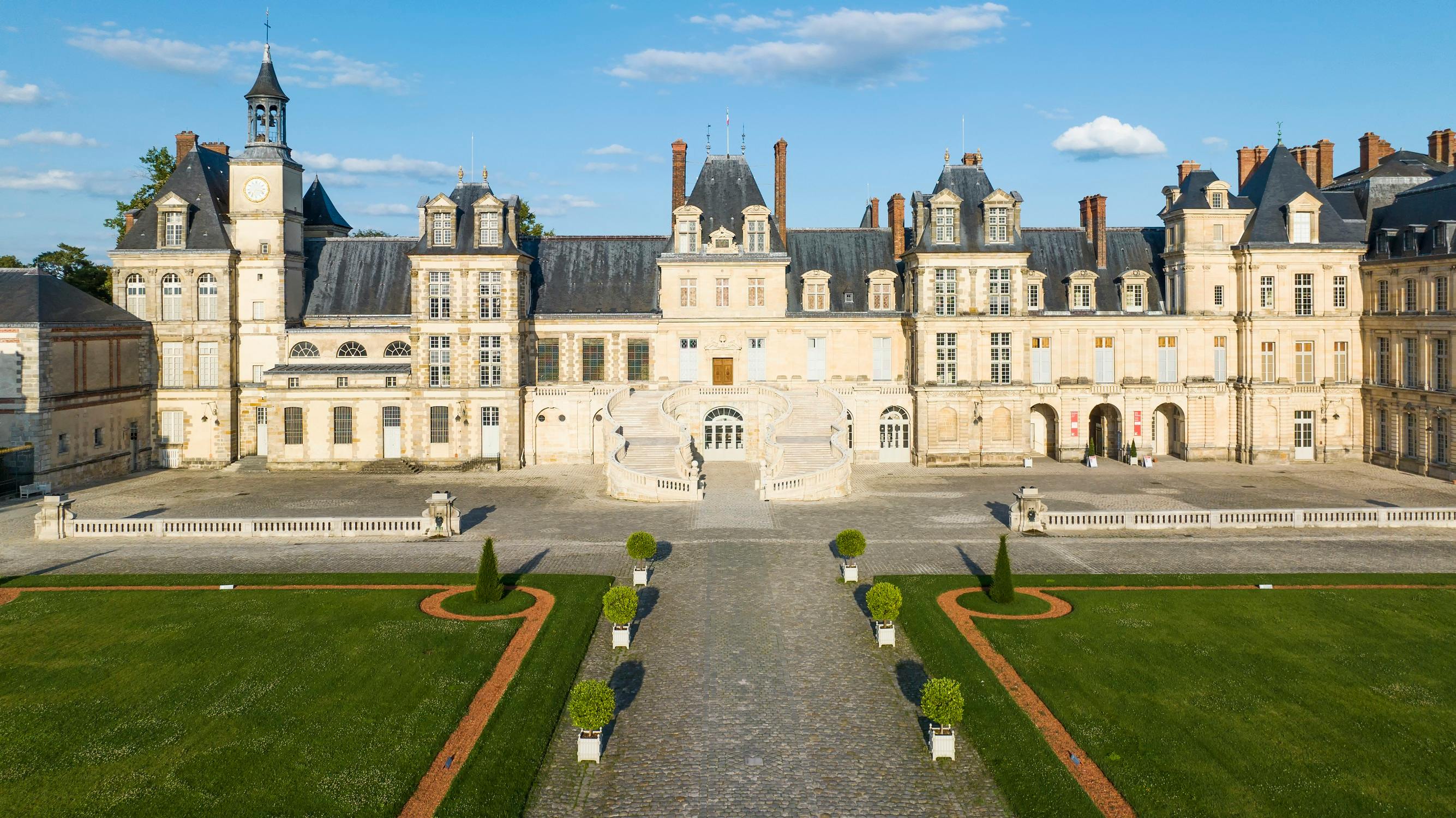 A grand historic building with multiple windows and chimneys, featuring a symmetrical garden and cobblestone pathway.