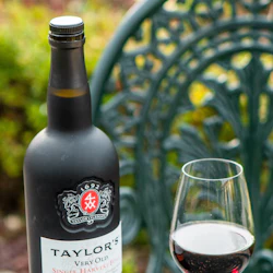 En flaska Taylors portvin och ett delvis fyllt vinglas på ett utomhusbord med en dekorativ metallstol i bakgrunden.