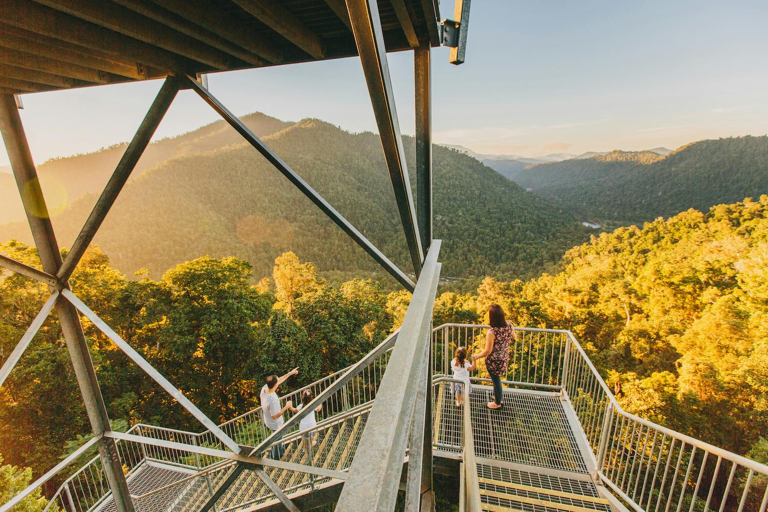 Three people standing on a metal viewing platform overlooking a forested mountain range during sunset.