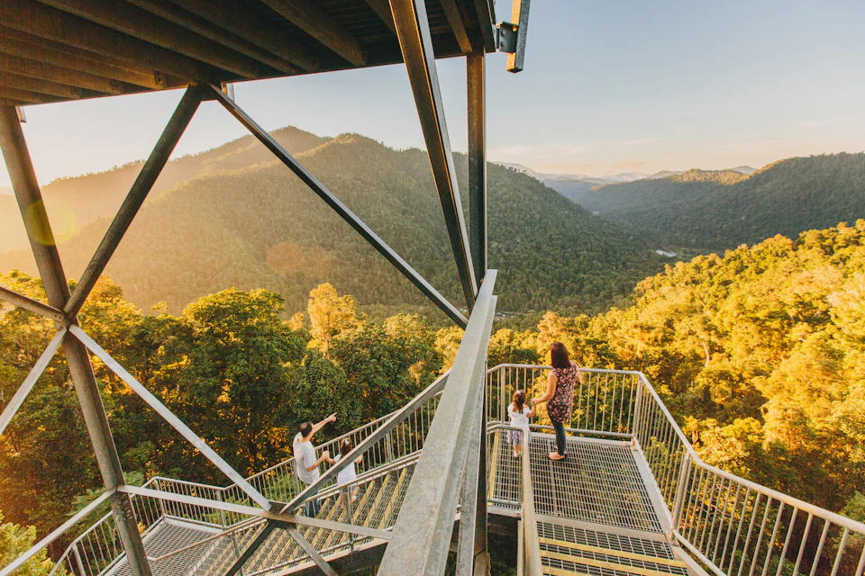 Three people standing on a metal viewing platform overlooking a forested mountain range during sunset.