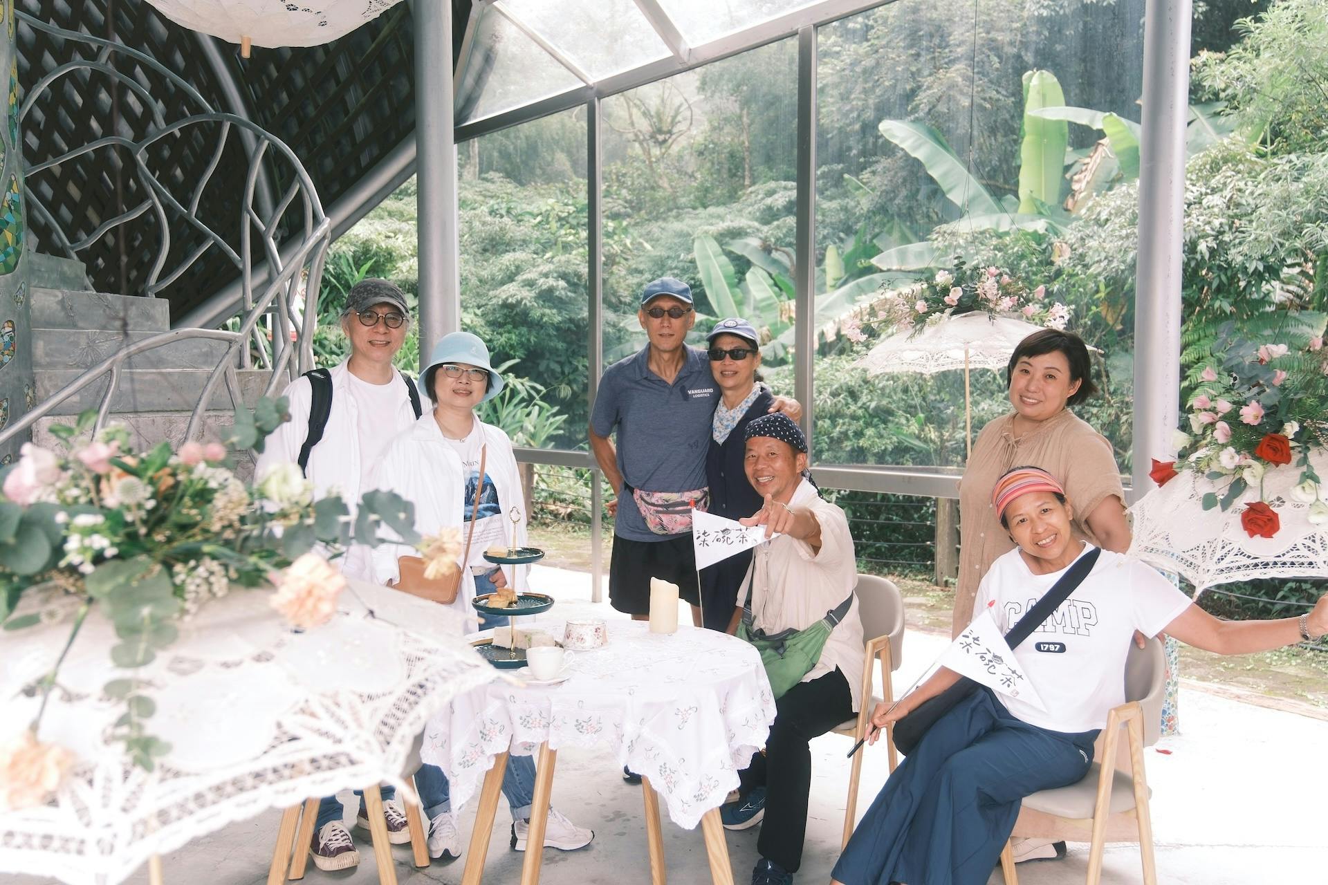 A group of people posing around a table with tea and decorations, set in an outdoor area with greenery visible through glass walls.