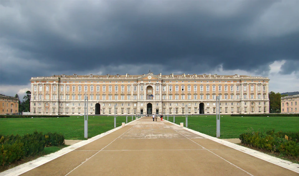 Large historical building with ornate facade, under dark clouds. People walking on a wide, paved path flanked by grassy areas.