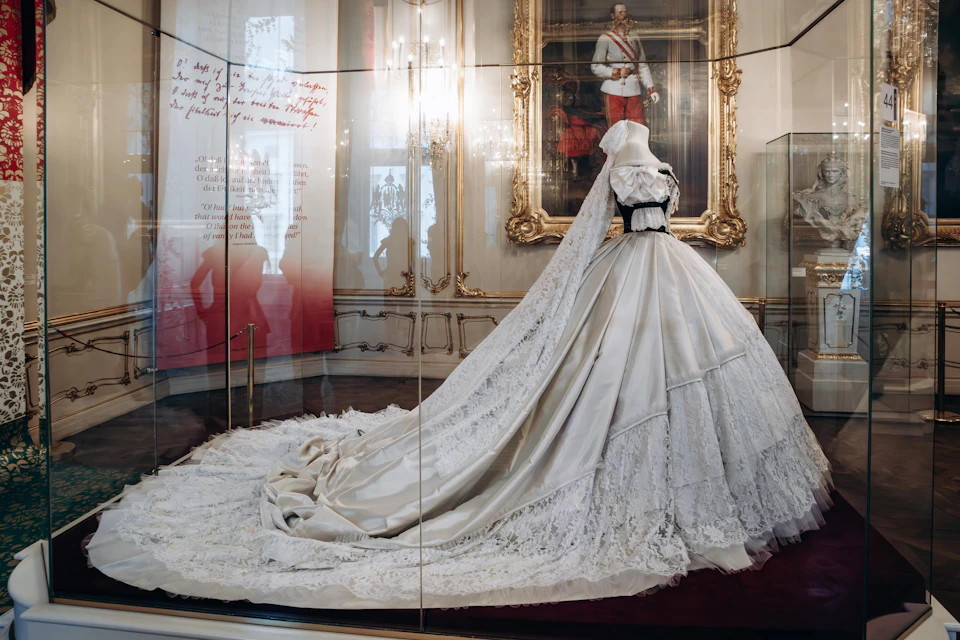 A white, ornate wedding gown with a long lace train displayed in a museum, with a painting of a regal figure in the background.