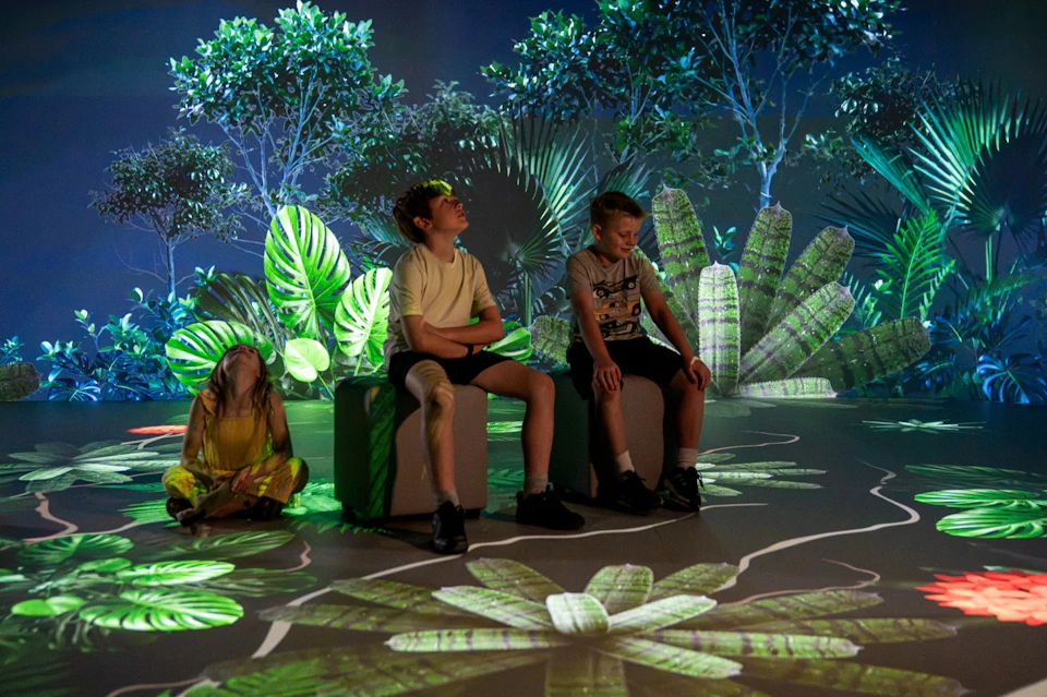 Two boys sit on cubes in a room with immersive tropical plant projections on the walls and floor.