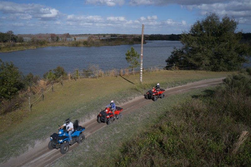 일부 흐린 하늘 아래 호숫가 흙길에서 ATV를 타고 있는 세 사람. 나무와 풀밭이 이 지역을 둘러싸고 있습니다.