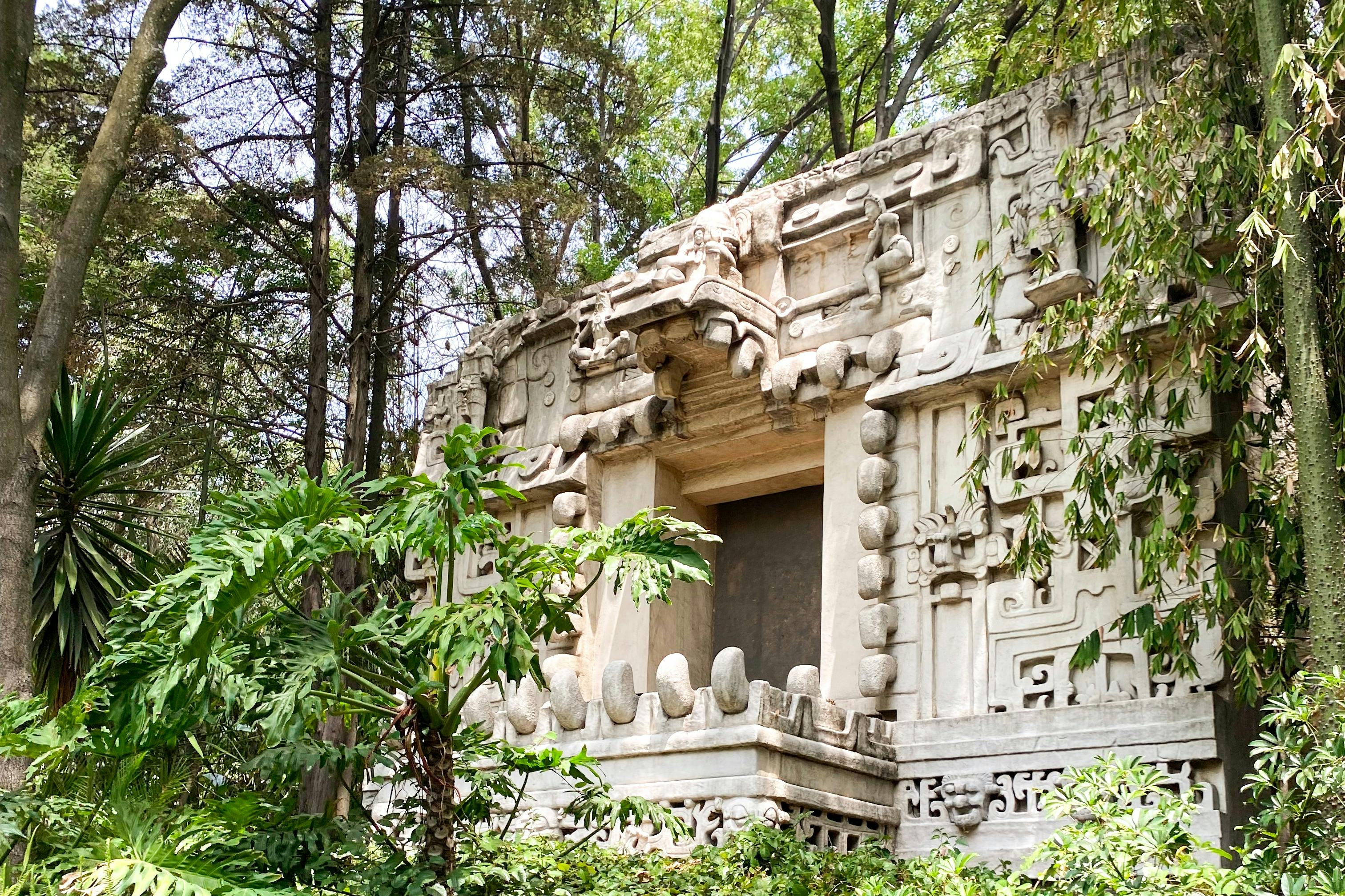 Struttura in pietra con intagli intricati circondata da una vegetazione lussureggiante e da alberi in un ambiente forestale.