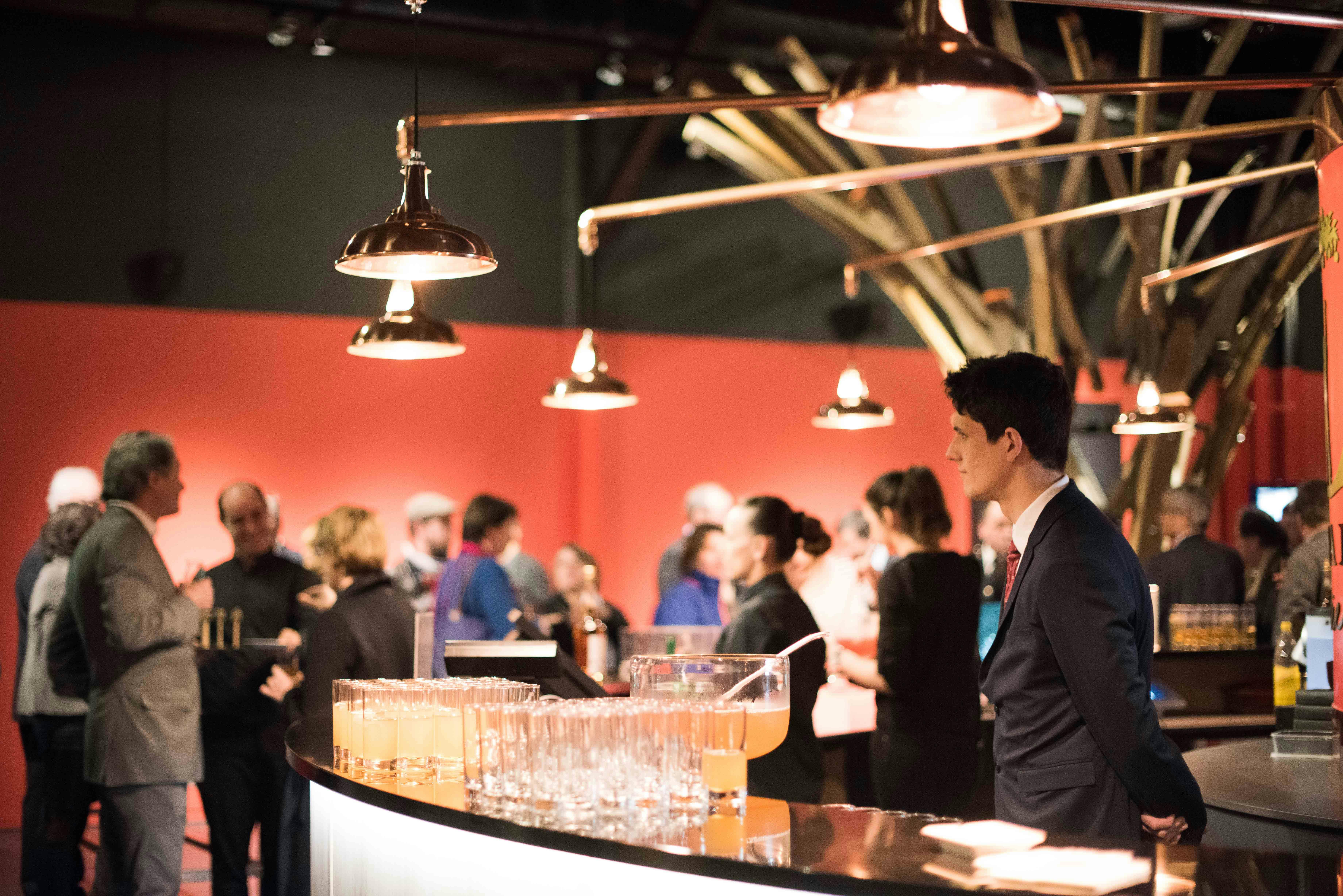 Un barman amb vestit es troba darrere d'un bar amb gerres de begudes i gots, amb gent socialitzant en un lloc ben il·luminat.