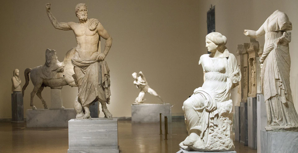 A museum gallery displaying ancient Greek marble statues, including seated and headless figures.