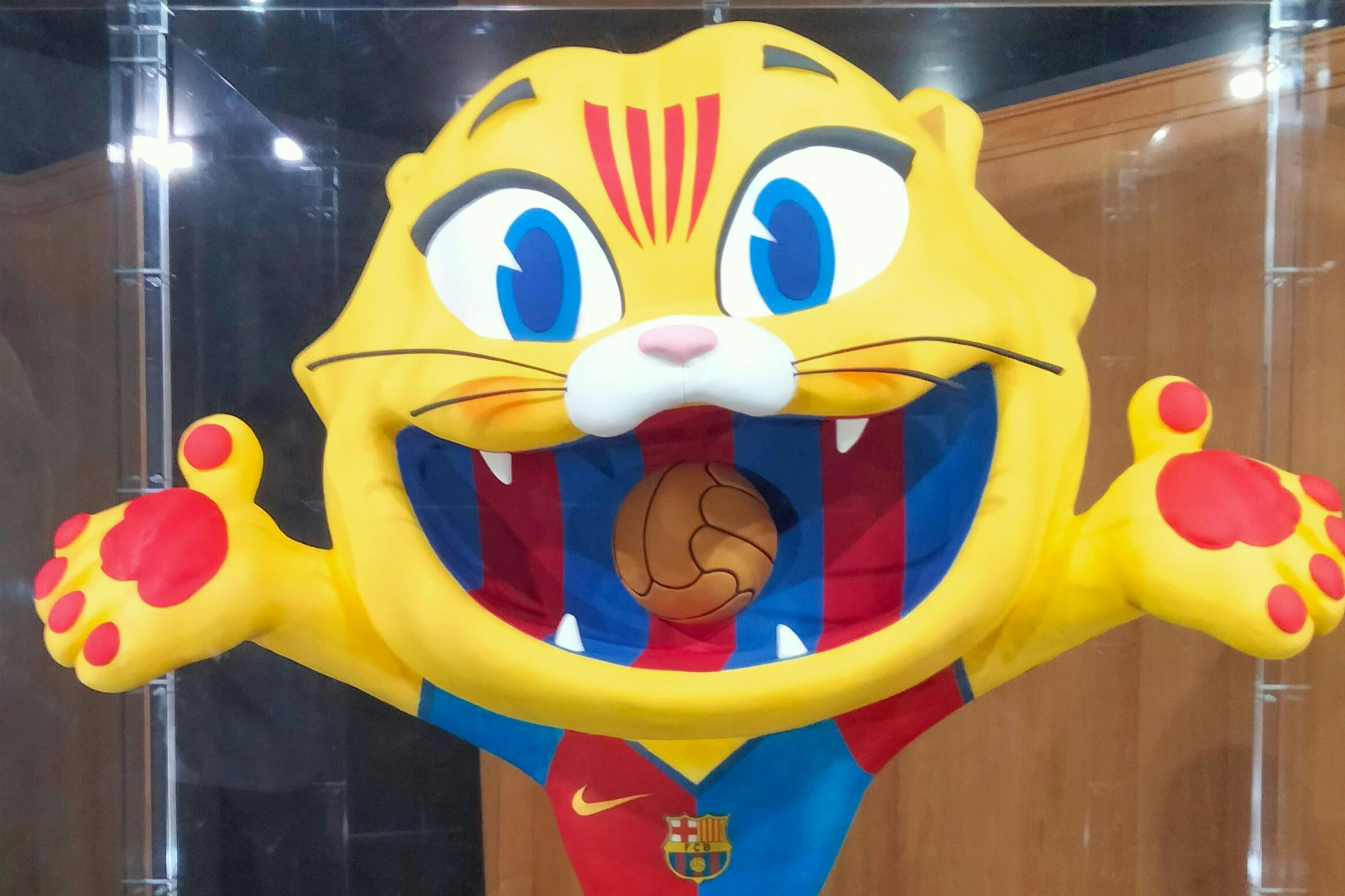 Une mascotte jaune caricaturale ressemblant à un chat à la bouche ouverte, portant un maillot bleu et rouge avec un ballon de football dans la bouche.