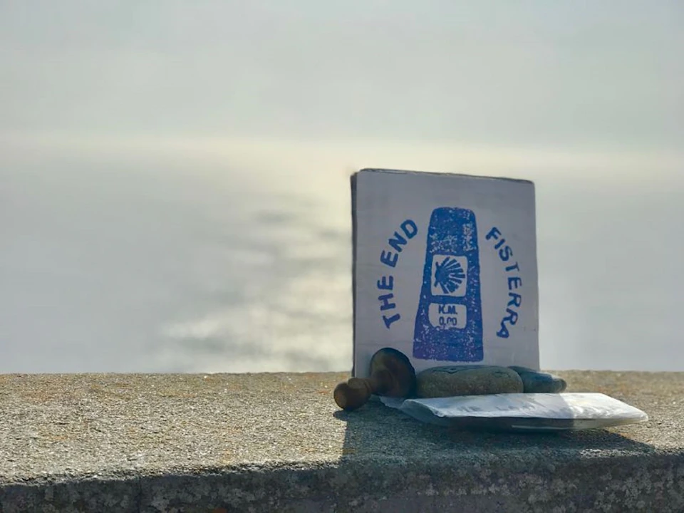 A sign marking "The End Fisterra KM 0,0" stands on a ledge, with ocean water and sunlight visible in the background.