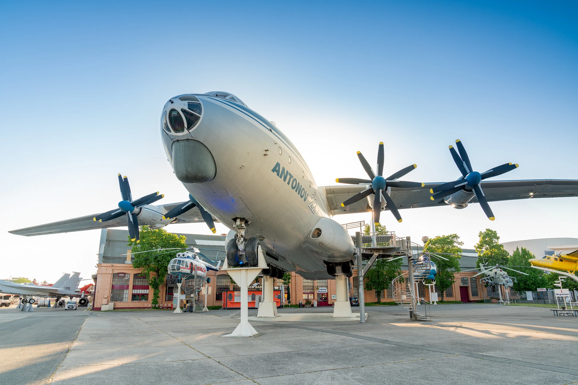 A large Antonov airplane displayed outdoors on stands, surrounded by smaller aircraft and buildings under a clear sky.