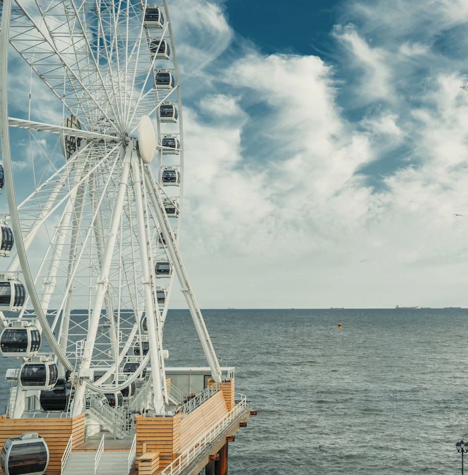 Large Ferris wheel with white gondolas next to a wooden pier, overlooking a calm ocean and a sky with scattered clouds.