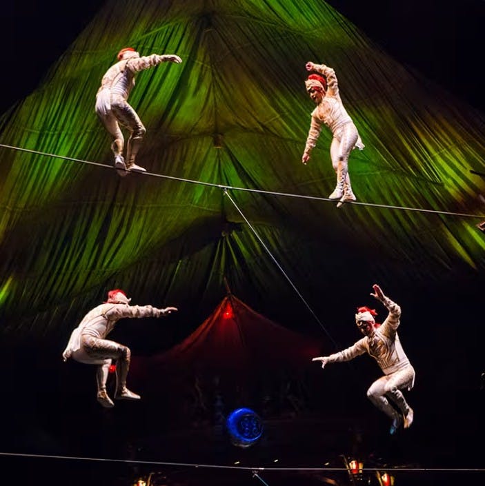Акробаты в белых костюмах и красных шляпах выступают на канате под зеленой цирковой палаткой.