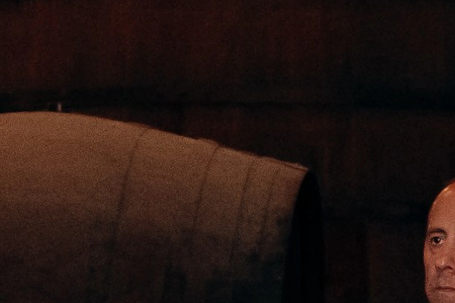 A group of five men in casual attire stand in a distillery or warehouse, surrounded by large wooden barrels, engaged in conversation.