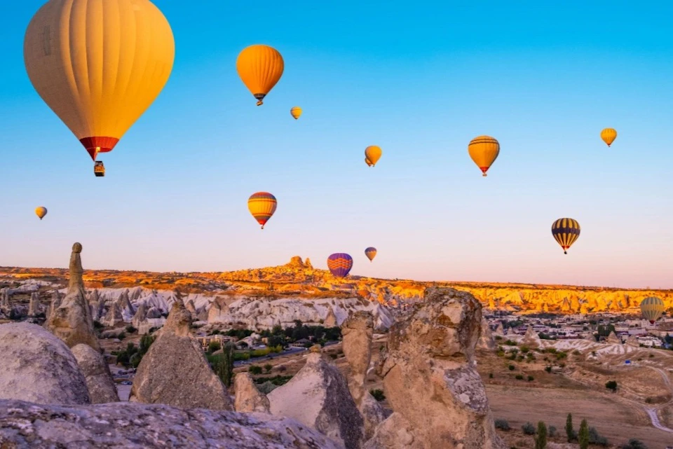 Hot air balloons of various colors floating over a rocky landscape at sunrise or sunset, with clear blue skies above.