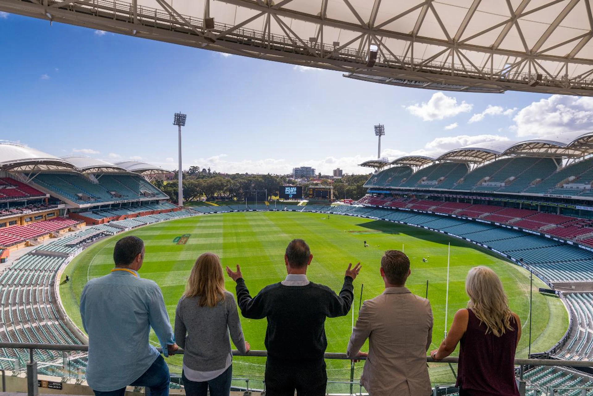 Five people stand in a stadium viewing area overlooking an empty football field with stands and a large screen in the distance.