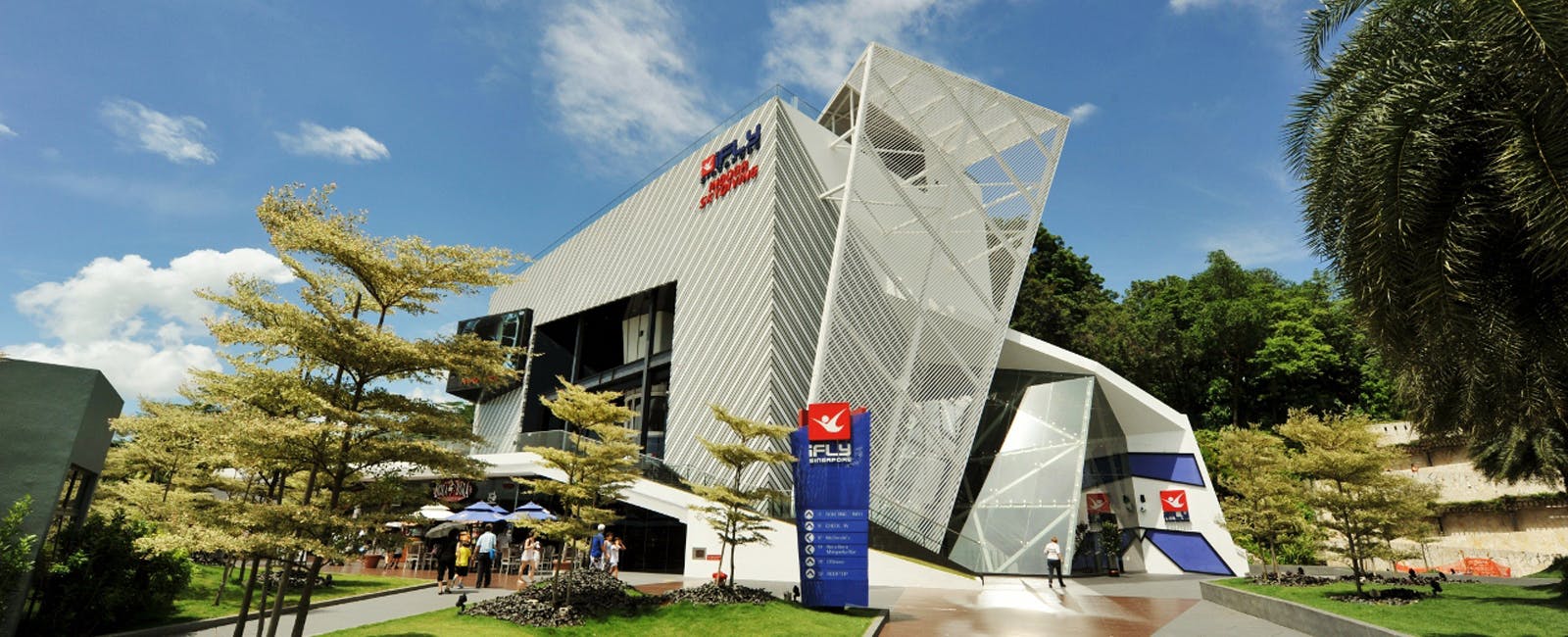 Modern building with a slanted design labeled &#34;iFly Singapore,&#34; surrounded by trees and people walking nearby on a sunny day.