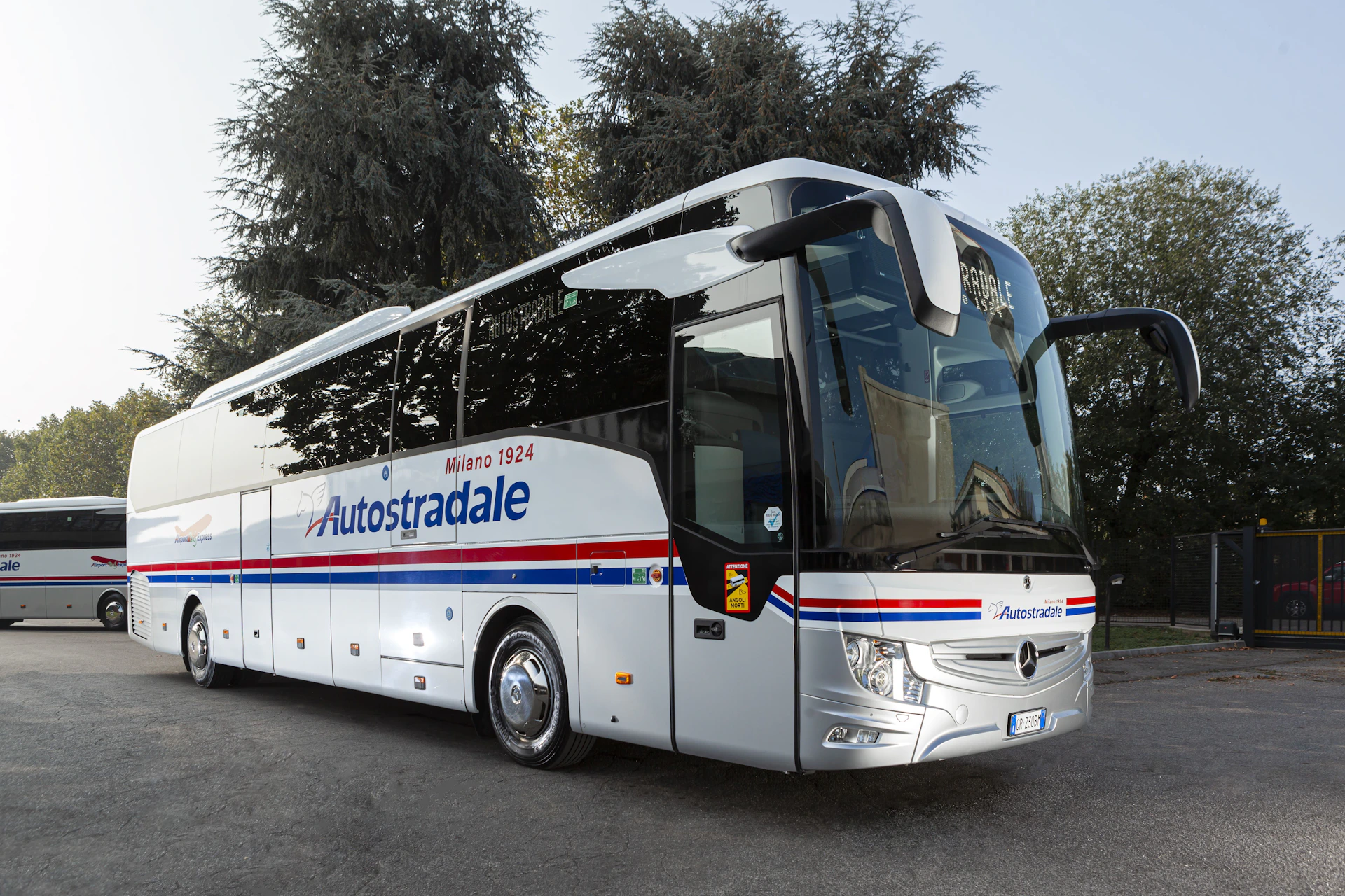 A white luxury coach bus with "Autostradale" branding, parked on a road with tall trees in the background.
