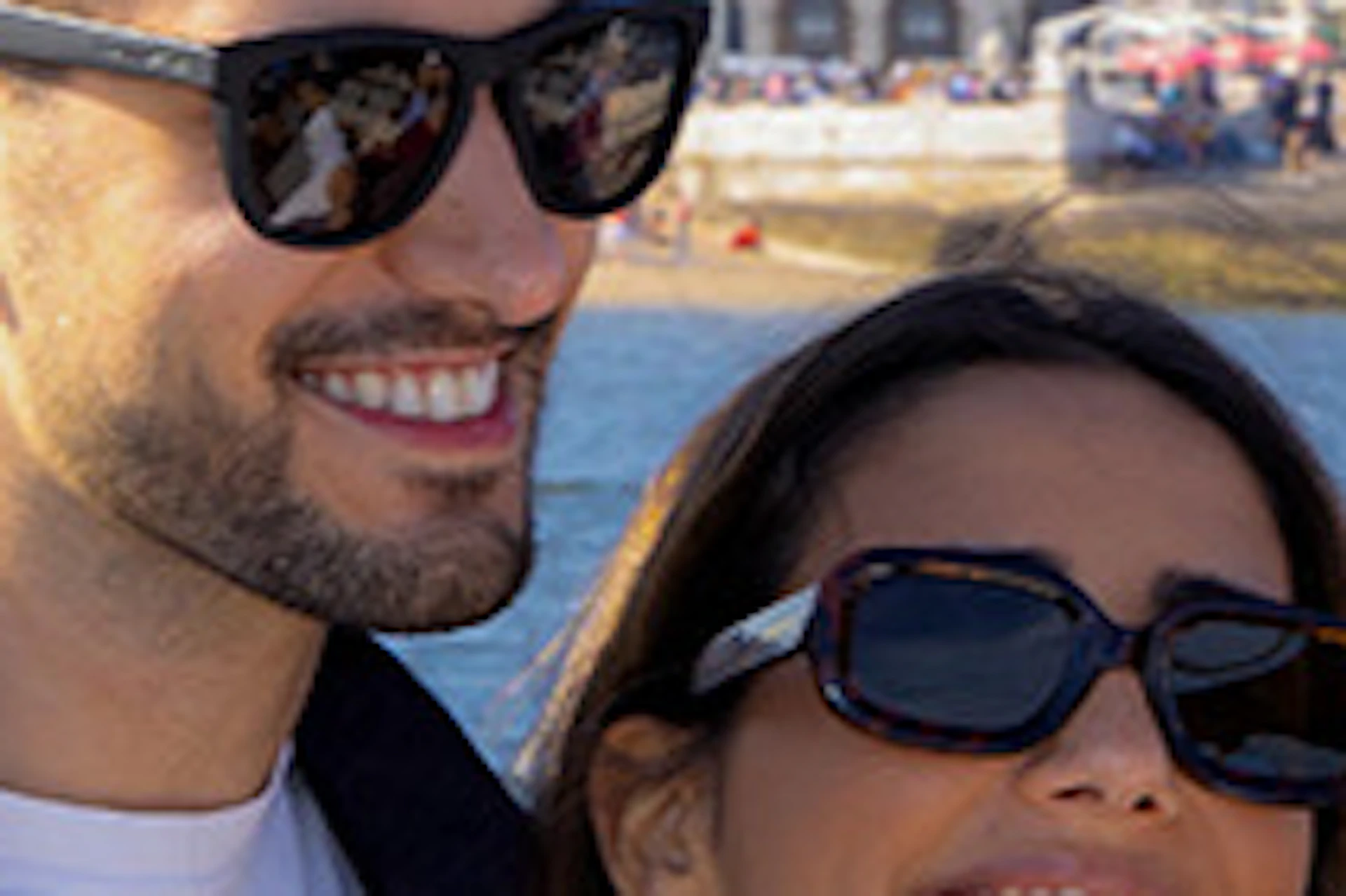 A smiling couple takes a selfie by a waterfront with a cityscape featuring a historic building and a sailboat in the background.