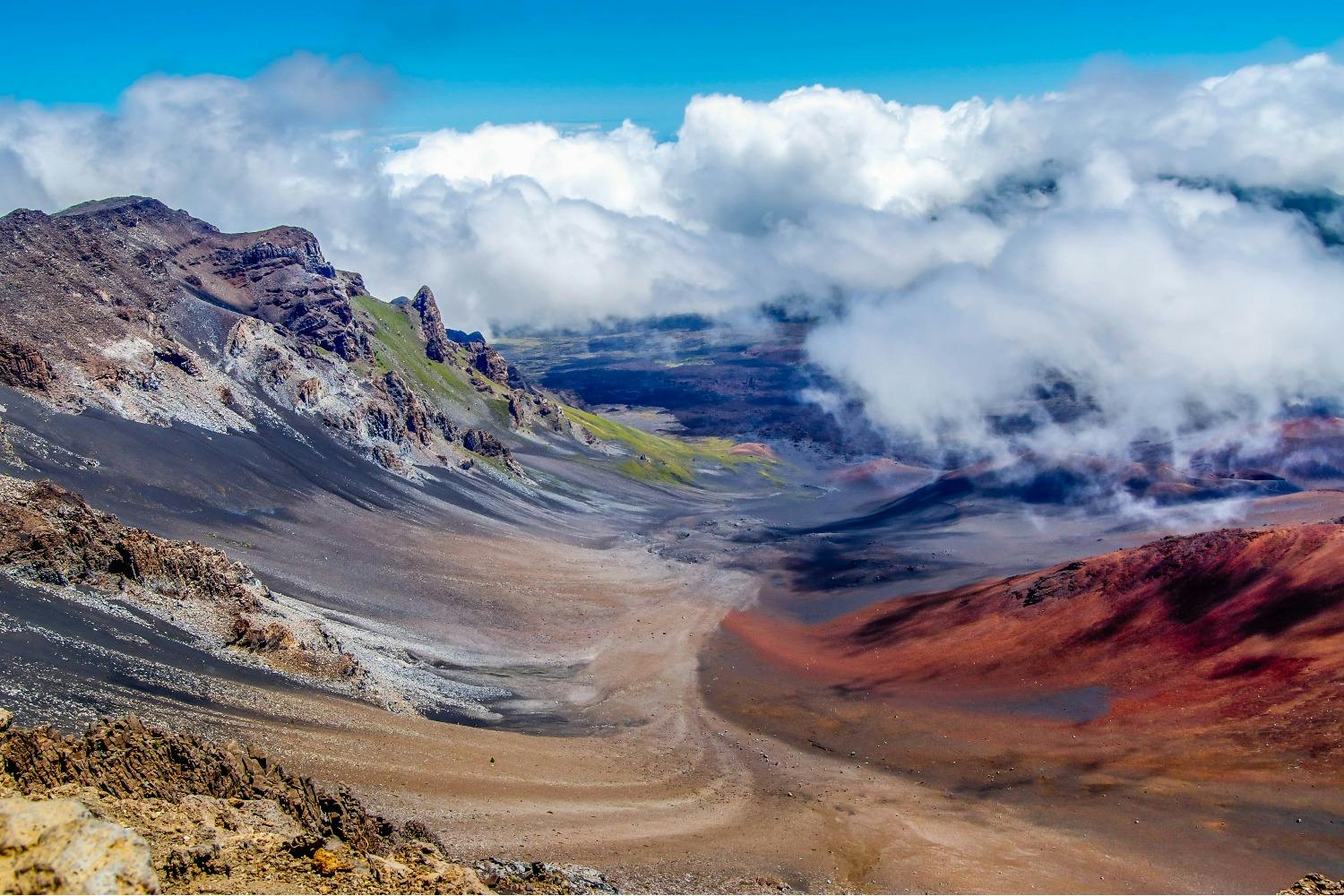 A colorful volcanic landscape featuring rocky terrain, red and brown hues, and clouds hovering over a crater.