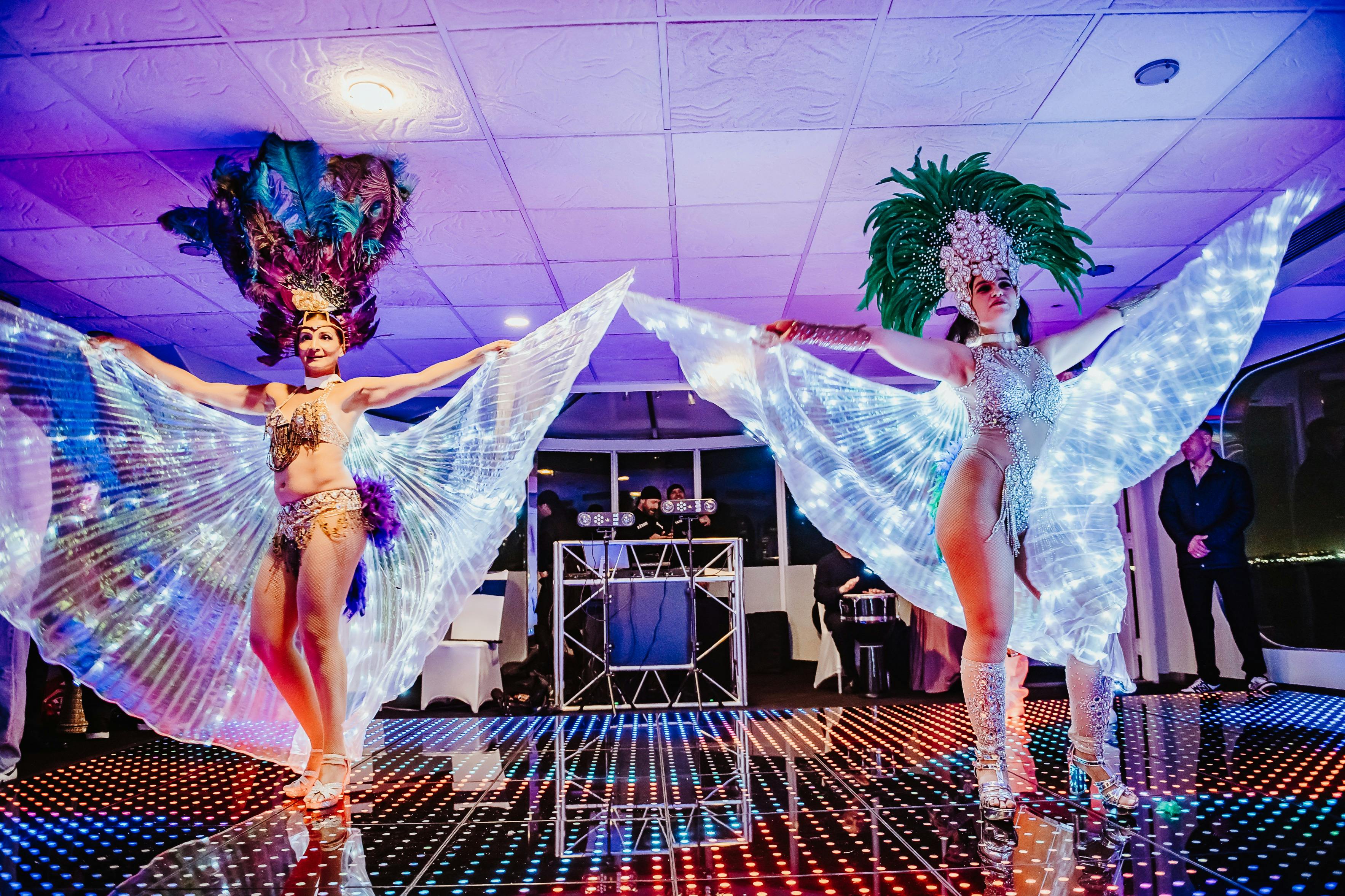 Účinkující v opeřené čelence a osvětlených křídlech tančí na barevném osvětleném pódiu v nočním klubu.