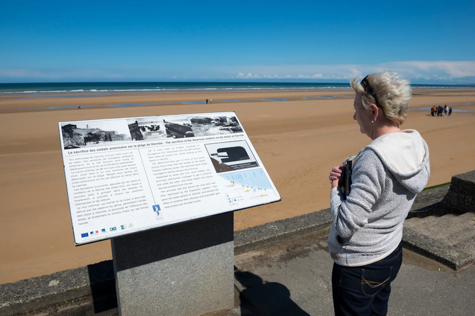 A person reads an informational sign by a sandy beach under a clear blue sky, with the ocean visible in the background.