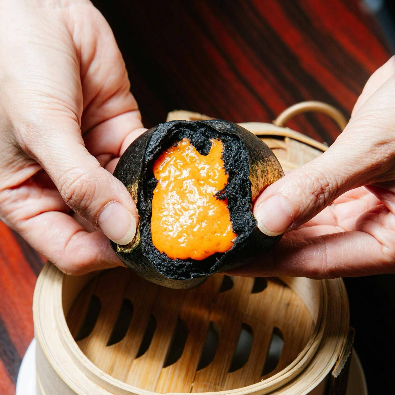 Hands opening a black steamed bun revealing orange filling, over a bamboo steamer on a wooden table.